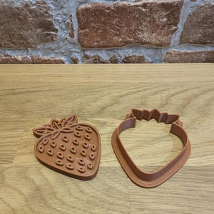 May include: Two brown strawberry-shaped cookie cutters. One is a solid shape with detailed seed impressions, and the other is a cutter outline. The cutters are made of plastic and sit on a wooden surface.