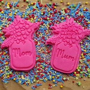 May include: Two pink cookies shaped like flower bouquets in a jar, with the word "Mom" on one and "Mum" on the other. The cookies are surrounded by colorful sprinkles on a wooden surface.