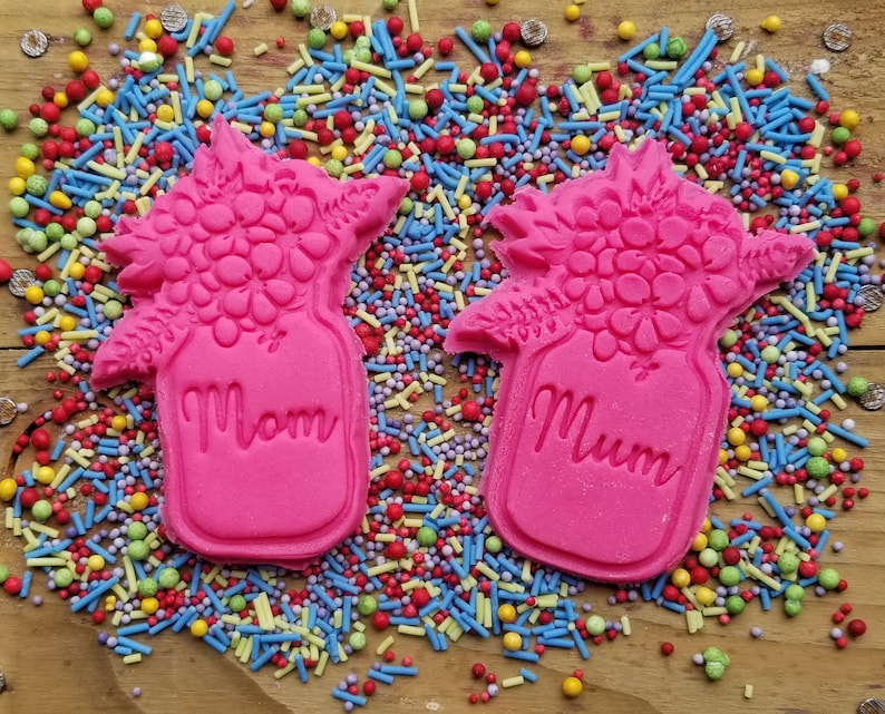 May include: Two pink cookies shaped like flower bouquets in a jar, with the word "Mom" on one and "Mum" on the other. The cookies are surrounded by colorful sprinkles on a wooden surface.