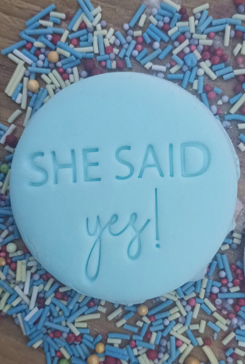 Może przedstawiać: Jasnoniebieski, okrągły fondant z wytłoczonymi słowami "SHE SAID yes!". Fondant otoczony jest kolorową posypką w kolorze niebieskim, białym, czerwonym i ż&oacute;łtym.