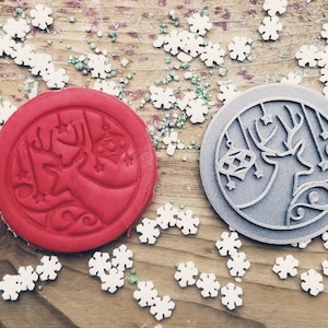 May include: A red circular cookie stamp with a reindeer design and a gray circular cookie stamp with a reindeer and star design. White snowflake and green sprinkles are scattered on a wooden surface.