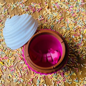 May include: A cupcake-shaped container with a white, ridged top and a brown and pink base. The container is open, revealing a pink interior and pink objects inside. The container sits on a wooden surface covered in colorful sprinkles.