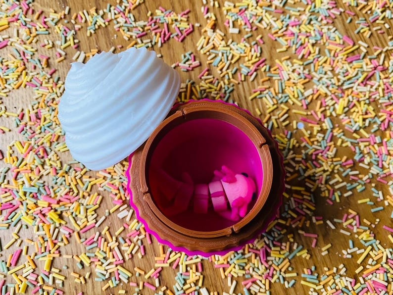 May include: A cupcake-shaped container with a white, ridged top and a brown and pink base. The container is open, revealing a pink interior and pink objects inside. The container sits on a wooden surface covered in colorful sprinkles.