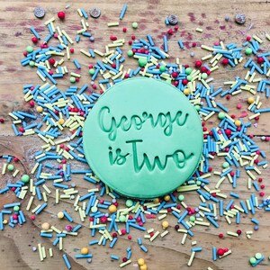 Op de afbeelding: Een ronde groene koek met de tekst "George is Two" in witte letters op een houten oppervlak bedekt met kleurrijke sprinkels.