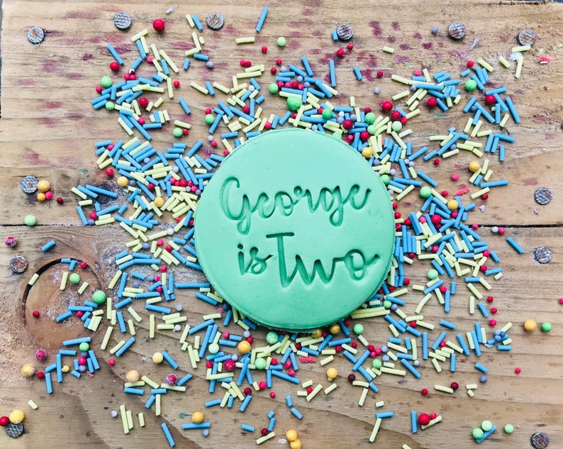 Op de afbeelding: Een ronde groene koek met de tekst "George is Two" in witte letters op een houten oppervlak bedekt met kleurrijke sprinkels.