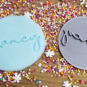 May include: Two round cookie cutters, one light blue with the name "Nancy" in cursive, and one gray with the name "Norman" in cursive.