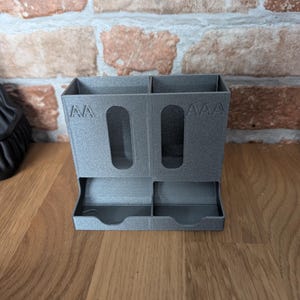 May include: A gray, 3D-printed desk organizer with two compartments and two small trays. The organizer has cut-out handles and the letters "AA" and "AAA" are printed on the front. The organizer is sitting on a wooden surface.