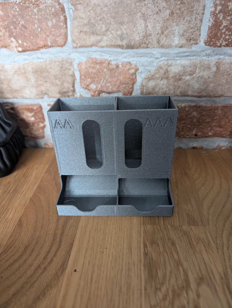 May include: A gray, 3D-printed desk organizer with two compartments and two small trays. The organizer has cut-out handles and the letters "AA" and "AAA" are printed on the front. The organizer is sitting on a wooden surface.