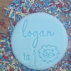 Puede incluir: Decoraci&oacute;n de pastel circular azul claro con el nombre "Logan", las palabras "is 1" y una ilustraci&oacute;n de le&oacute;n. La decoraci&oacute;n est&aacute; rodeada de chispas de colores sobre una superficie de madera.