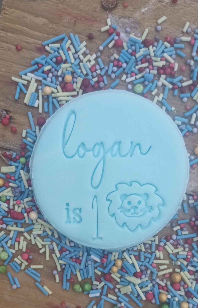 Puede incluir: Decoraci&oacute;n de pastel circular azul claro con el nombre "Logan", las palabras "is 1" y una ilustraci&oacute;n de le&oacute;n. La decoraci&oacute;n est&aacute; rodeada de chispas de colores sobre una superficie de madera.