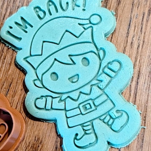 May include: A light blue cookie cutter in the shape of an elf with the text "I'M BACK!" The elf is wearing a hat, belt, and shoes. The cookie cutter is on a wooden surface.