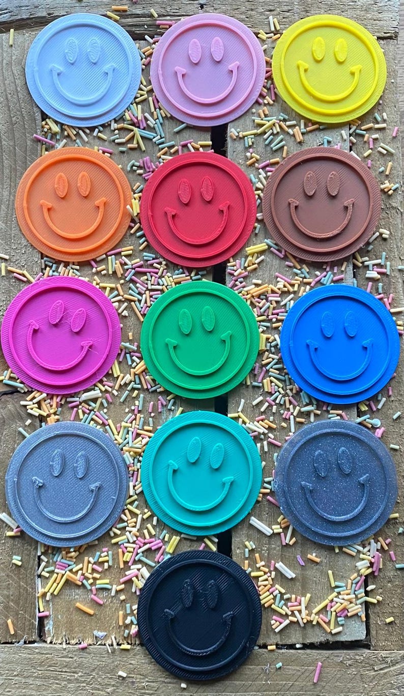 May include: Twelve round, colorful cookie cutters in the shape of a smiley face. The cutters are arranged in a circle on a wooden surface with colorful sprinkles.