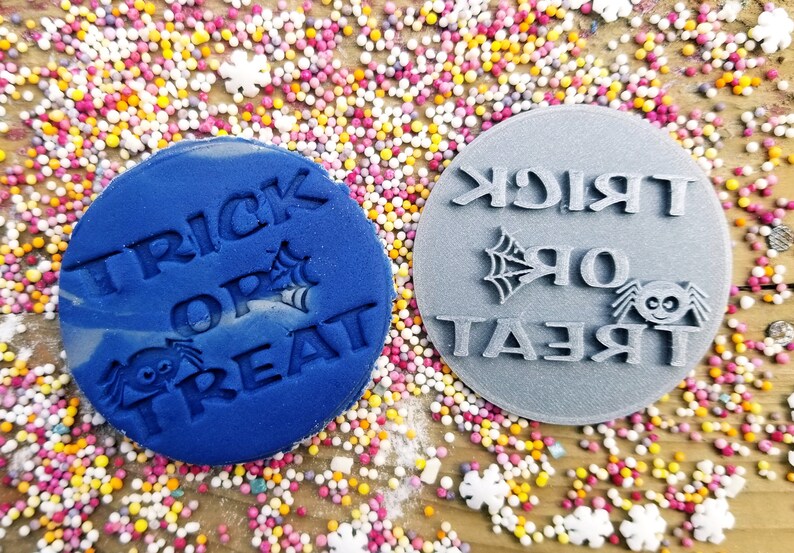 Puede incluir: Dos sellos circulares para galletas con tem&aacute;tica de Halloween. Uno es azul con las palabras "TRICK OR TREAT", una ara&ntilde;a y una telara&ntilde;a. El otro es gris con el mismo dise&ntilde;o. El fondo est&aacute; cubierto de chispas de colores.