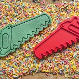 May include: Two cookie cutters shaped like saws, one green and one red, rest on a bed of colorful sprinkles. The cutters have a handle and a jagged blade design. The sprinkles are a mix of yellow, pink, blue, and white.