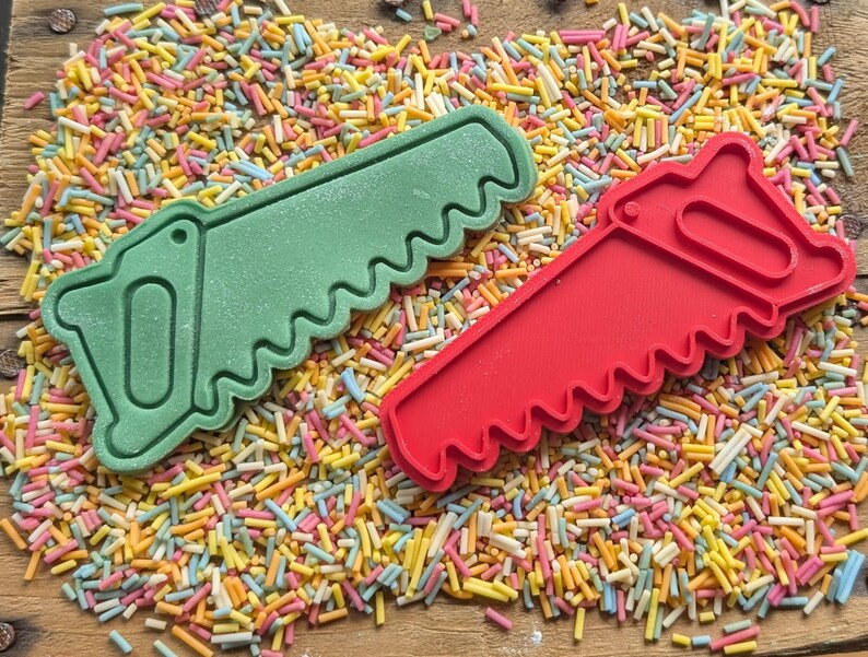 May include: Two cookie cutters shaped like saws, one green and one red, rest on a bed of colorful sprinkles. The cutters have a handle and a jagged blade design. The sprinkles are a mix of yellow, pink, blue, and white.