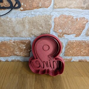 May include: A red plastic cookie cutter in the shape of the number "9" with the word "nine" written in cursive script. The cutter is on a wooden surface with a brick wall in the background.