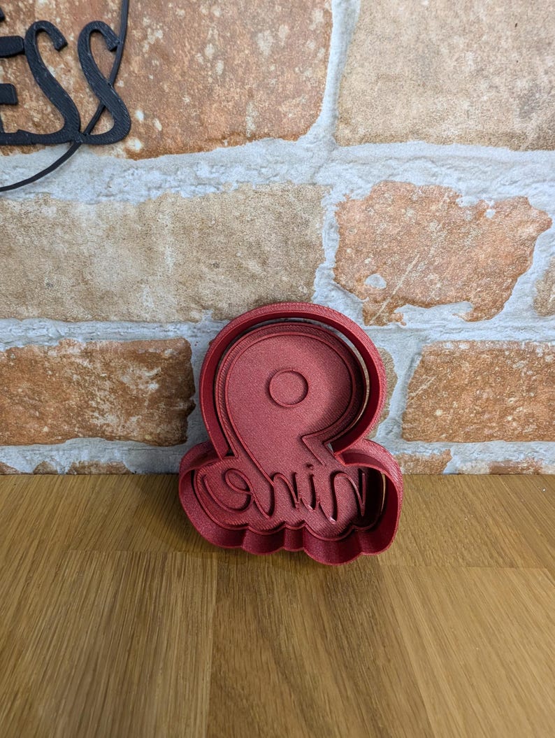 May include: A red plastic cookie cutter in the shape of the number "9" with the word "nine" written in cursive script. The cutter is on a wooden surface with a brick wall in the background.