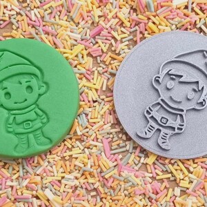 May include: Two round cookie cutters, one green and one gray, with a raised design of a cartoon elf wearing a hat.