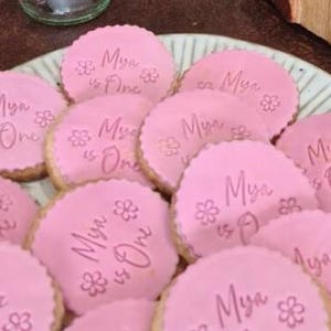 May include: Pink sugar cookies decorated with white icing and the text "Mya is One" in pink lettering.