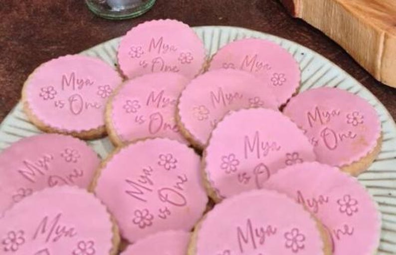 May include: Pink sugar cookies decorated with white icing and the text "Mya is One" in pink lettering.