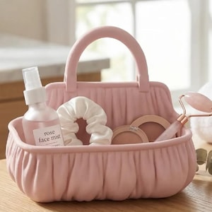 May include: A pink fabric basket with a handle, containing beauty items. Inside are a rose face mist, a white scrunchie, two compacts, and a rose quartz face roller. The basket sits on a wooden surface.