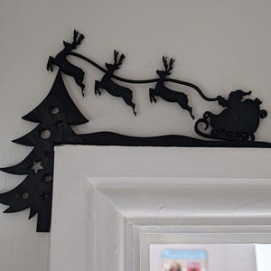 May include: Black metal Christmas decoration featuring Santa Claus in a sleigh pulled by reindeer, with a Christmas tree on the left. The decoration is placed above a white door frame, creating a festive holiday scene.