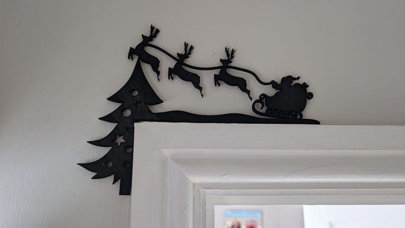 May include: Black metal Christmas decoration featuring Santa Claus in a sleigh pulled by reindeer, with a Christmas tree on the left. The decoration is placed above a white door frame, creating a festive holiday scene.