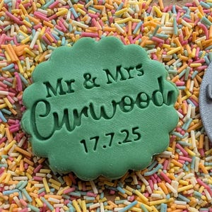 Puede incluir: Un sello verde con borde festoneado con el texto "Mr & Mrs Curwood 17.7.25" est&aacute; rodeado de chispas de colores. Tambi&eacute;n es visible un sello gris circular con texto en cursiva, lo que sugiere un tema de boda.