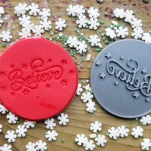 May include: Two circular cookie stamps, one red and one gray, each imprinted with the word "Believe" in a script font, surrounded by small stars. White snowflake sprinkles and green sugar crystals are scattered around the stamps on a wooden surface.