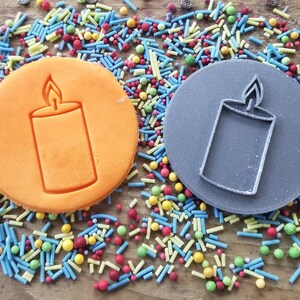 May include: Two circular cookie cutters, one orange and one grey, each featuring a candle design. The cutters are surrounded by colourful sprinkles on a wooden surface. The candle design is a rectangular shape with a flame.