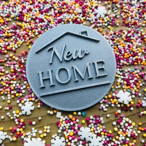 May include: A gray, circular emblem with the words "New Home" and a house outline. The emblem is surrounded by colorful sprinkles and snowflake-shaped decorations, creating a festive and celebratory scene.