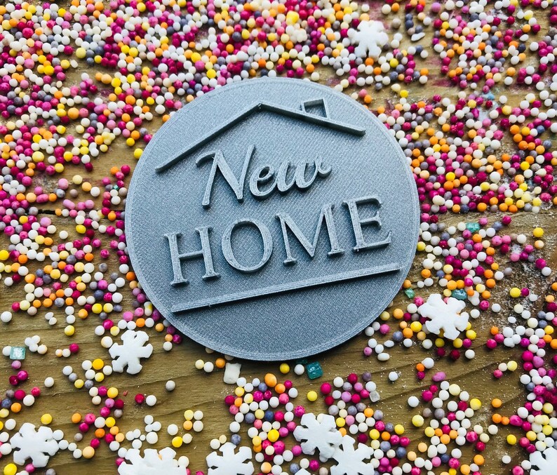 May include: A gray, circular emblem with the words "New Home" and a house outline. The emblem is surrounded by colorful sprinkles and snowflake-shaped decorations, creating a festive and celebratory scene.