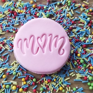 May include: A round, light pink cookie stamp with the word "MOM" in a cursive font, with a heart replacing the "O". The stamp is surrounded by colorful sprinkles on a wooden surface.