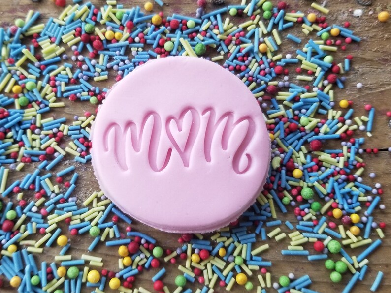 May include: A round, light pink cookie stamp with the word "MOM" in a cursive font, with a heart replacing the "O". The stamp is surrounded by colorful sprinkles on a wooden surface.