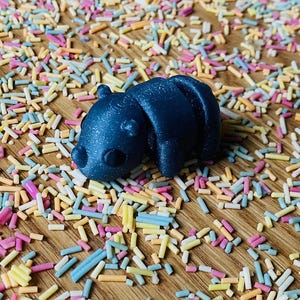 May include: A dark blue 3D printed animal figurine, possibly a bear or a pig, lying on a surface covered in colorful sprinkles.