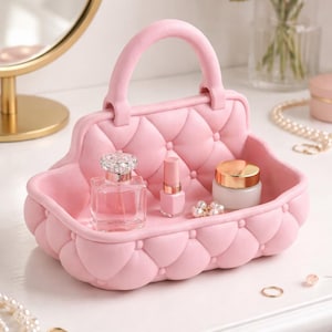 May include: A pink, quilted handbag-shaped tray with a handle, holding perfume, lipstick, and a jar of cream. The tray is decorated with a diamond pattern and sits on a white surface, with jewelry and flowers in the background.