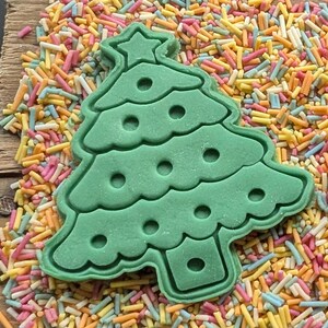 Puede incluir: Un cortador de galletas verde en forma de &aacute;rbol de Navidad con una estrella en la parte superior, rodeado de chispas de colores. El cortador tiene un dise&ntilde;o detallado con recortes circulares, perfecto para hornear en Navidad.