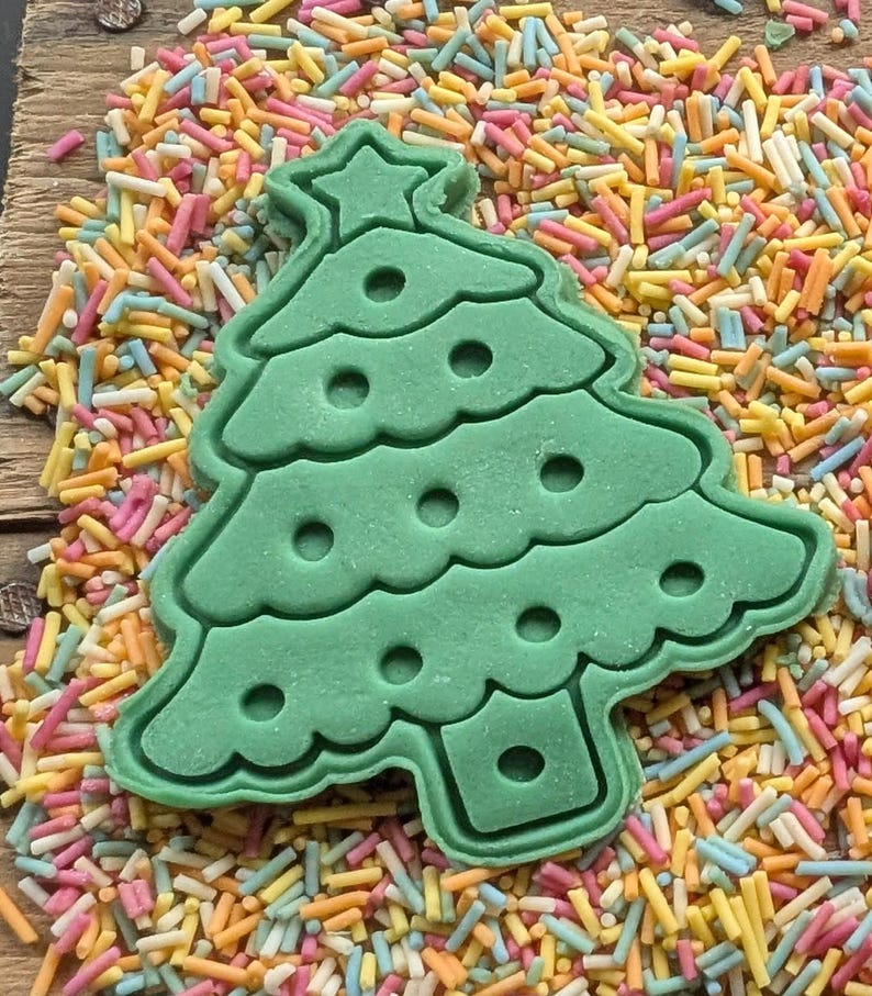 Puede incluir: Un cortador de galletas verde en forma de &aacute;rbol de Navidad con una estrella en la parte superior, rodeado de chispas de colores. El cortador tiene un dise&ntilde;o detallado con recortes circulares, perfecto para hornear en Navidad.