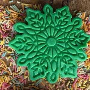 May include: A bright green snowflake-shaped cookie cutter with intricate floral details. The cutter is surrounded by colorful sprinkles on a wooden surface. Ideal for baking holiday treats.