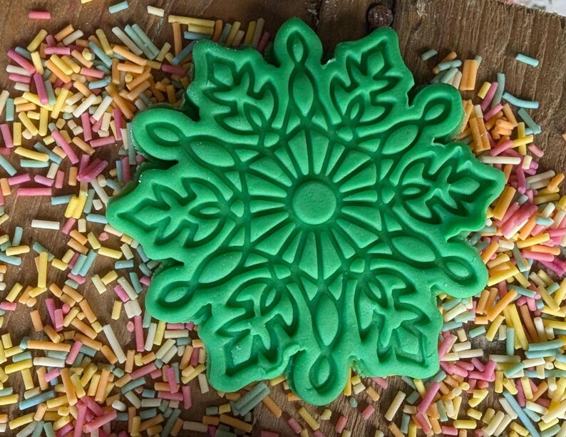 May include: A bright green snowflake-shaped cookie cutter with intricate floral details. The cutter is surrounded by colorful sprinkles on a wooden surface. Ideal for baking holiday treats.