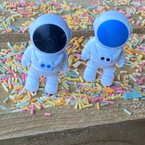 K&ouml;nnte beinhalten: Zwei wei&szlig;e Astronautenfiguren mit blauen und schwarzen Visieren, die auf einer mit bunten Streuseln bedeckten Holzoberfl&auml;che stehen.