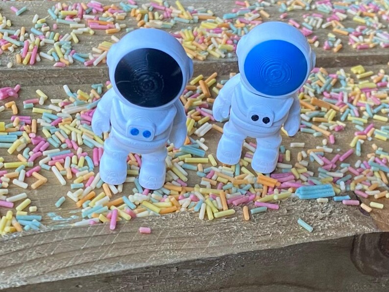 K&ouml;nnte beinhalten: Zwei wei&szlig;e Astronautenfiguren mit blauen und schwarzen Visieren, die auf einer mit bunten Streuseln bedeckten Holzoberfl&auml;che stehen.