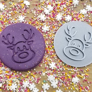 May include: Two round, purple and grey silicone cookie stamps with a reindeer design. The stamps are on a wooden surface with colourful sprinkles.