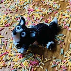 May include: A black and white toy dog figurine with a playful expression, lying on a surface covered in colorful sprinkles.
