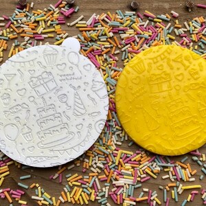 May include: Two round, embossed cake toppers, one white and one yellow, with a birthday theme. The designs include cakes, gifts, balloons, and the words "Happy Birthday". Colorful sprinkles surround the toppers on a wooden surface.