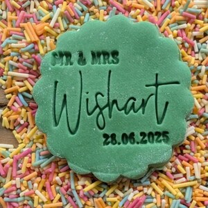 Op de afbeelding: Een groene koekje met geschulpte randen met de tekst "SIR & MRS Wishart 28.06.2025" is omringd door kleurrijke hagelslag. De koekje is felgroen van kleur en de tekst is in het oppervlak gegraveerd.