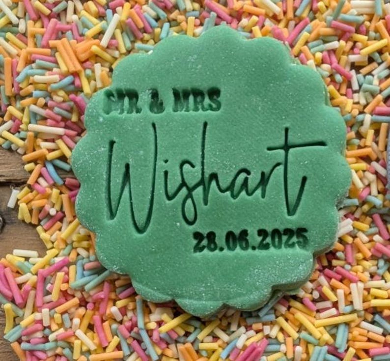 Op de afbeelding: Een groene koekje met geschulpte randen met de tekst "SIR & MRS Wishart 28.06.2025" is omringd door kleurrijke hagelslag. De koekje is felgroen van kleur en de tekst is in het oppervlak gegraveerd.