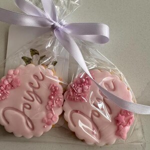 May include: Two pink frosted cookies in clear cellophane, tied with a lavender ribbon. The cookies have scalloped edges and are decorated with the name "Joyce" in cursive and floral accents. One cookie is visible with the name "Joyce".