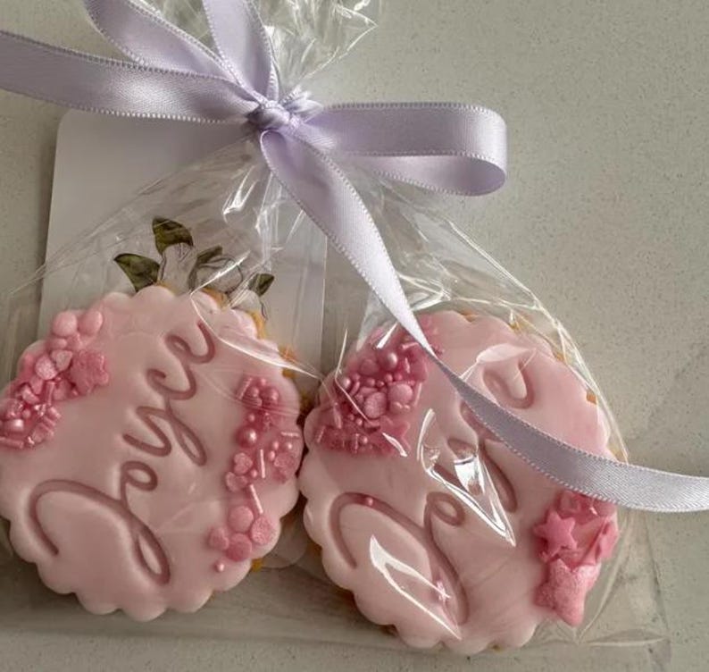 May include: Two pink frosted cookies in clear cellophane, tied with a lavender ribbon. The cookies have scalloped edges and are decorated with the name "Joyce" in cursive and floral accents. One cookie is visible with the name "Joyce".