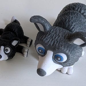 May include: Two husky dog figurines. One is lying down, the other is upright. Both have black and white markings, with blue eyes. The figurines are made of a textured material.